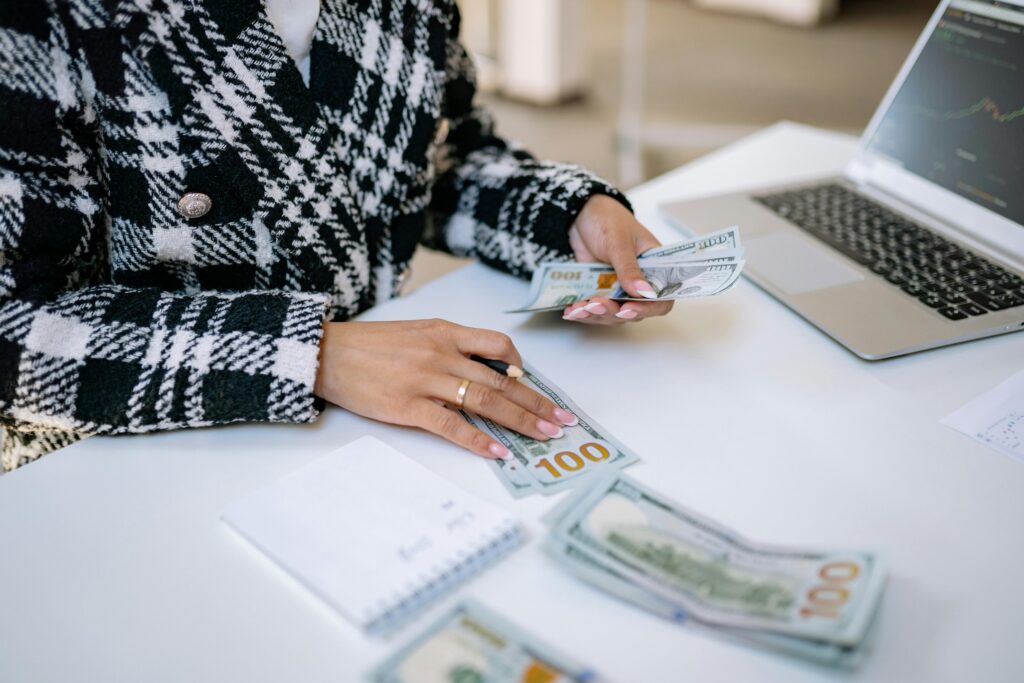 A woman shown from the neck down wearing a black and white checked coat with an open laptop counting money 