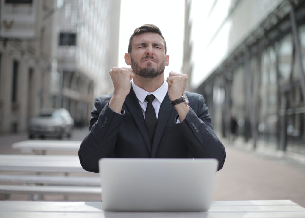 A young, business suit wearing man at a table with a laptop celebrating a possible victory with his hands clasped in front of him and eyes closed