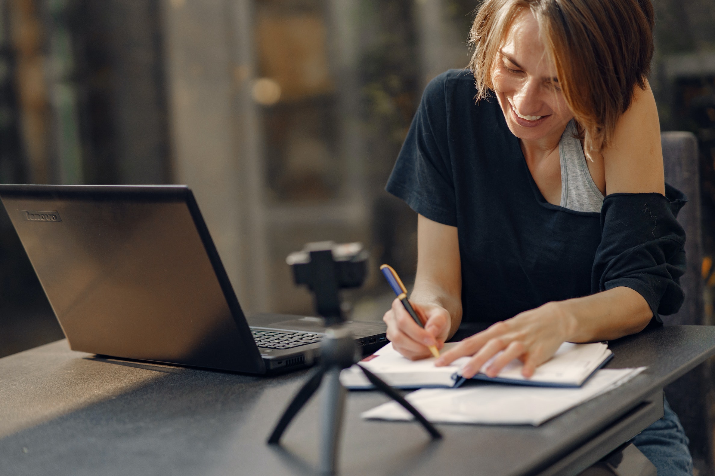 Dark haired woman writing notes in front of a laptop and a small video camera