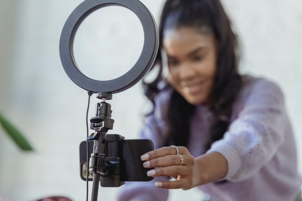 A young, dark-haired woman preparing to broadcast herself using her smartphone attached to a stand that is holding a ring light