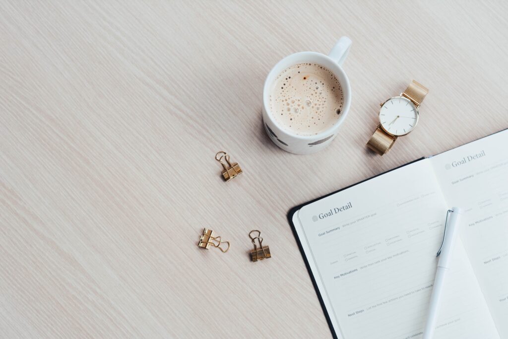 An overhead view of a light colored desktop with an organizer, a cup of hot beverage, a watch and three paper clips 