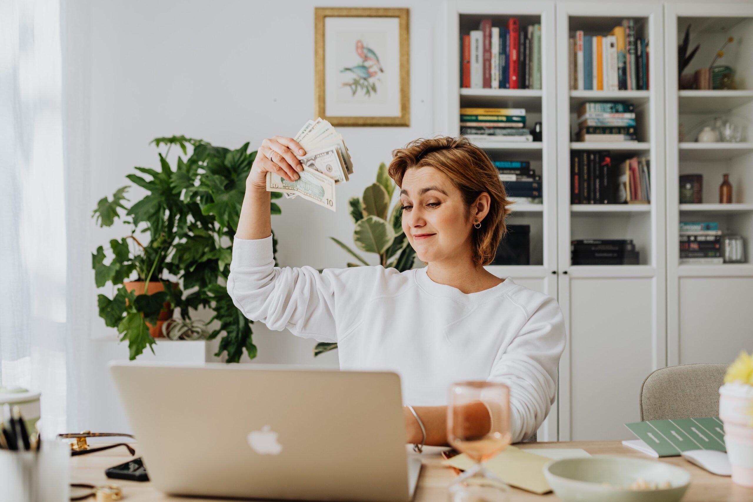 woman in while sweater holding money happily while looking at a laptop