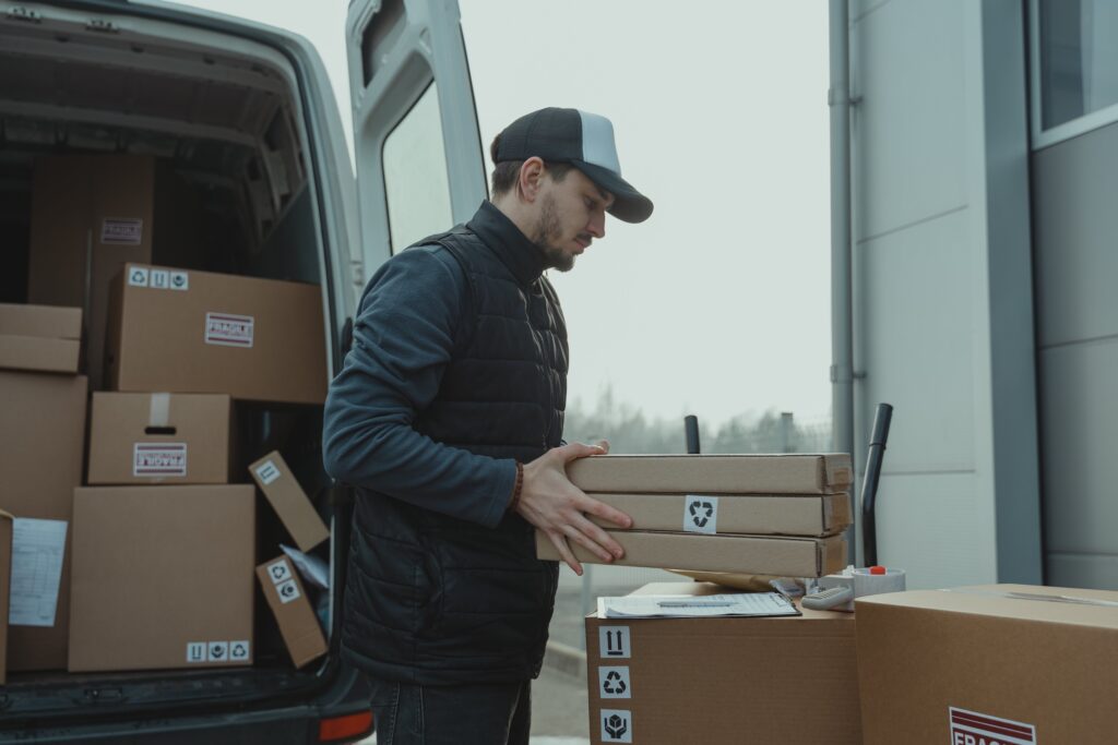 A male delivery driver holding three small cardboard boxes outside of the back of his open delivery van