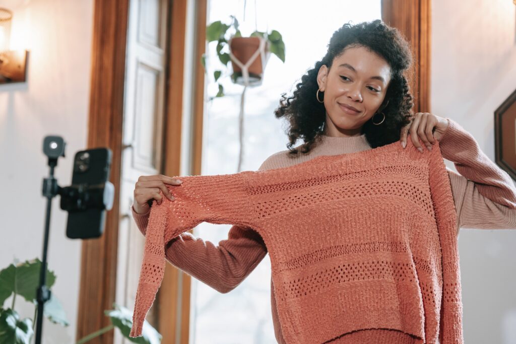 A young, dark haired influencer showing off a peach colored sweater to her audience over a smartphone broadcast