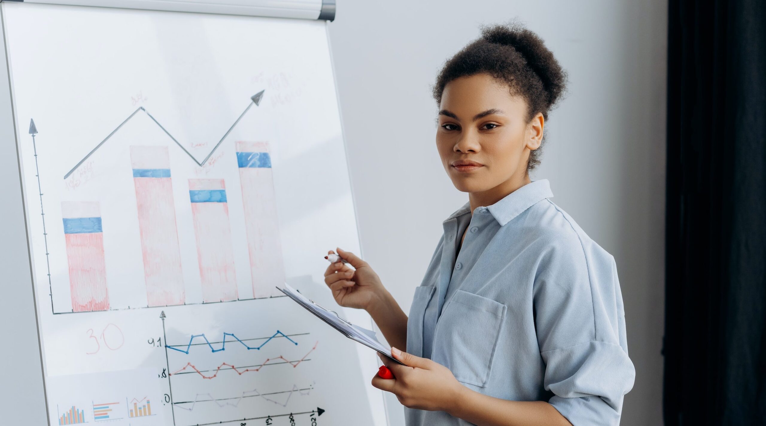 dark haired young woman holding a clipboard in front of a whiteboard