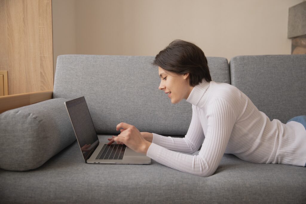 A woman laying on her couch is typing on a laptop. She is leaving a positive customer review on an Amazon product. 