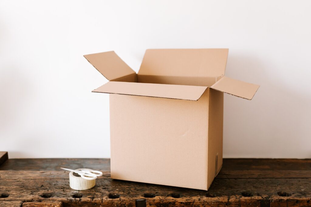 An empty cardboard box on a woooden table accompanied by a roll of tape and scissors