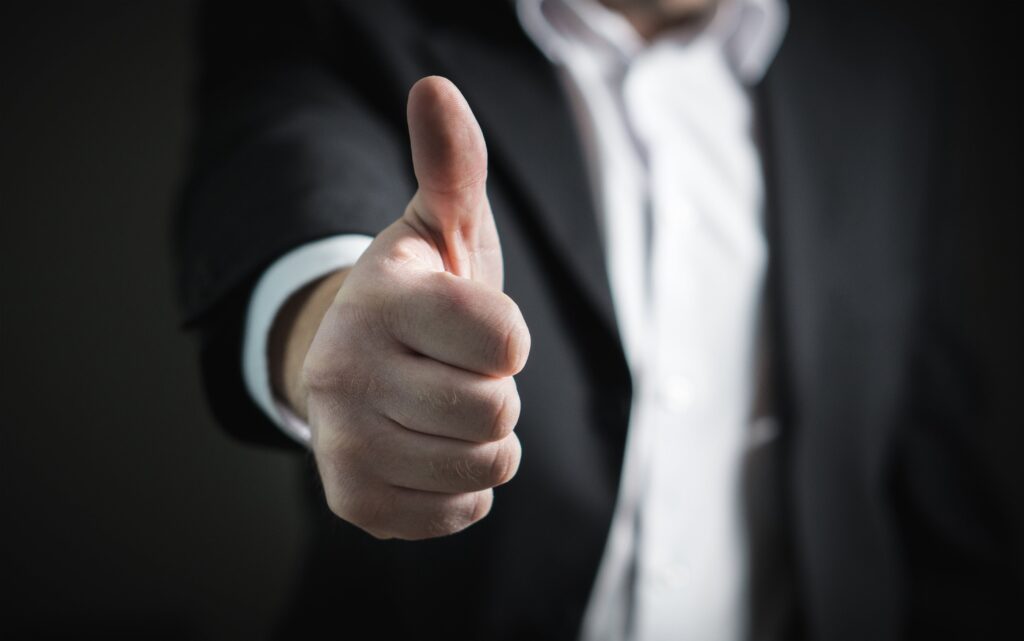 A man in a black business suit with his right hand outstretched, thumb raised 