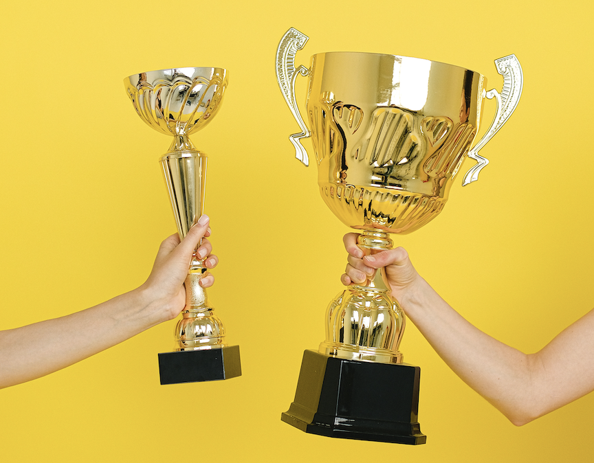 Two hands holding two gold trophies against a yellow background